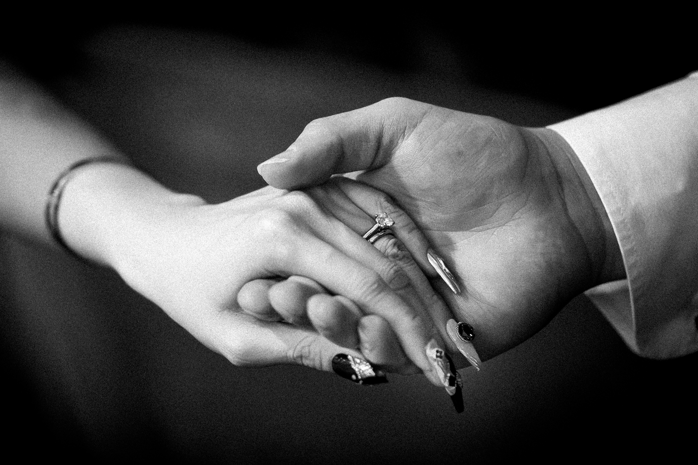 A black and white image of two hands gently holding each other evokes the elegance of a Duke Chapel wedding. The hand on the left, adorned with a ring and long decorative nails, is held by the hand on the right, which is wearing a shirt. The image conveys a timeless sense of connection and support.