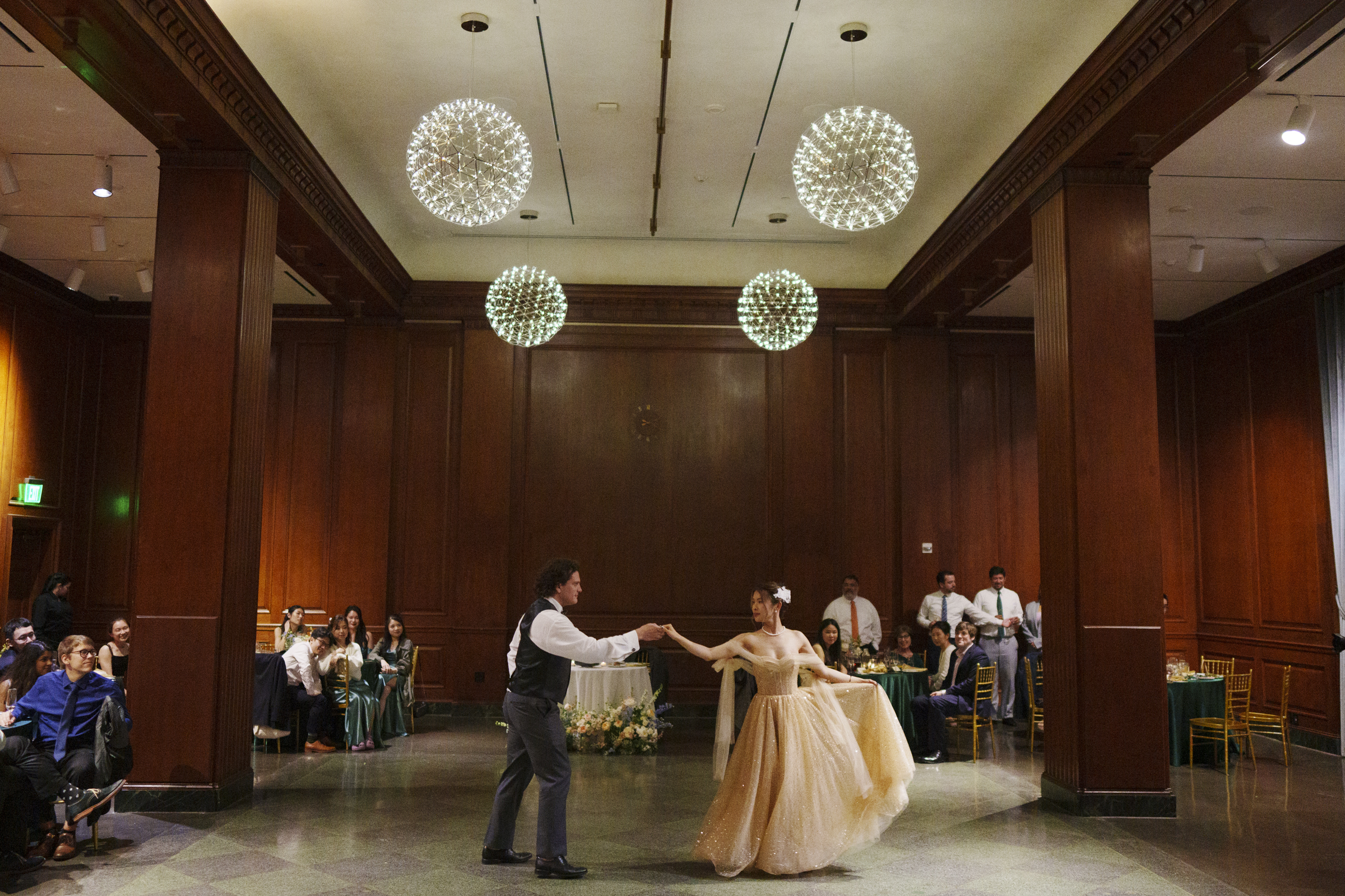 A couple dances together in an elegant ballroom with wood-paneled walls reminiscent of a Duke Chapel wedding. The woman wears a flowing gown, and the man is in a suit. Guests are seated at tables, watching the performance as sparkling chandeliers hang from the ceiling.