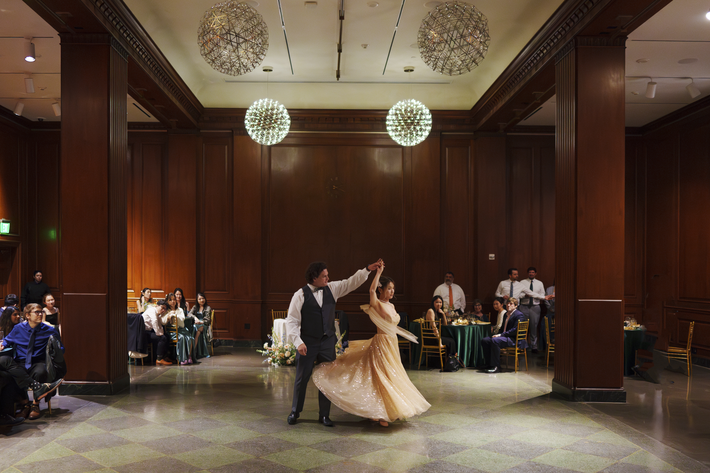 A couple elegantly dances in a wood-paneled ballroom, reminiscent of a Duke Chapel wedding. The woman wears a flowing gold dress, and the man is in formal attire. Guests watch from tables adorned with green tablecloths, while round chandeliers illuminate the room.