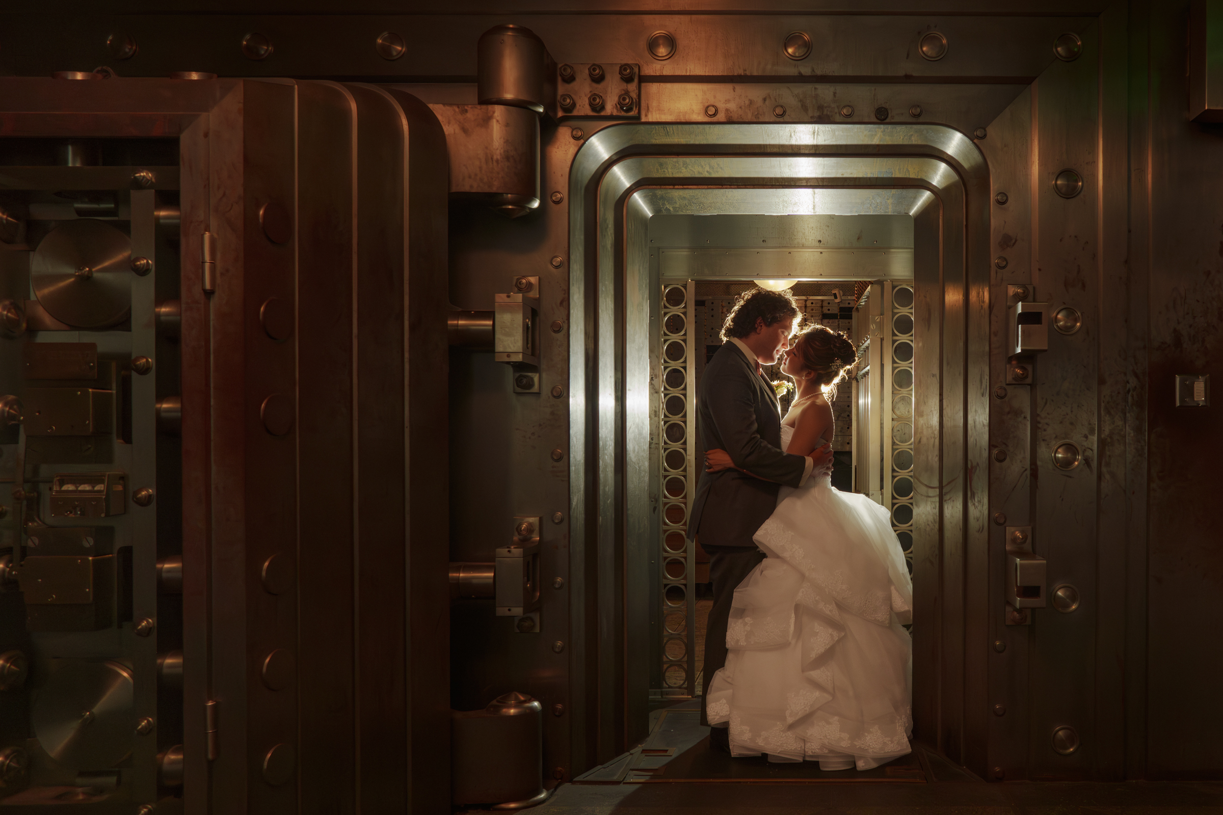 In a dimly lit, ornate metal vault reminiscent of a Duke Chapel wedding, a bride in her white gown and her groom in a suit share an embrace. Light casts a warm glow around them, highlighting the intricate details of the vault door.