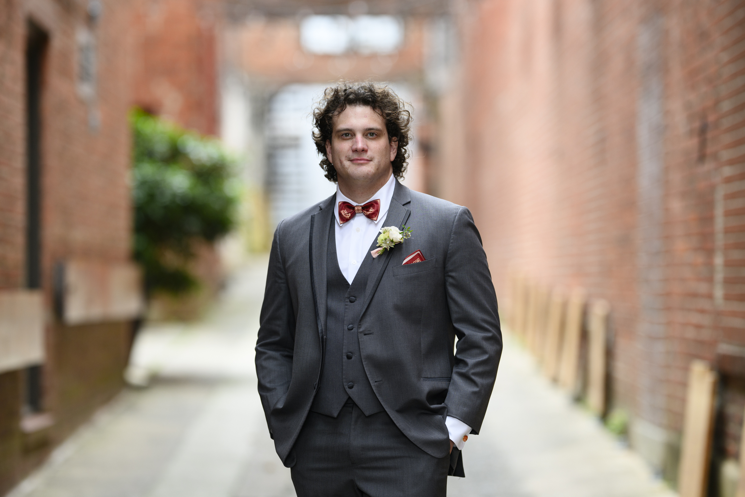 In the narrow alleyway, a man stands poised in a gray three-piece suit with a red bow tie and white shirt. Curly-haired and adorned with a boutonniere, he seems ready for a Duke Chapel wedding against the backdrop of brick walls and blurred greenery.