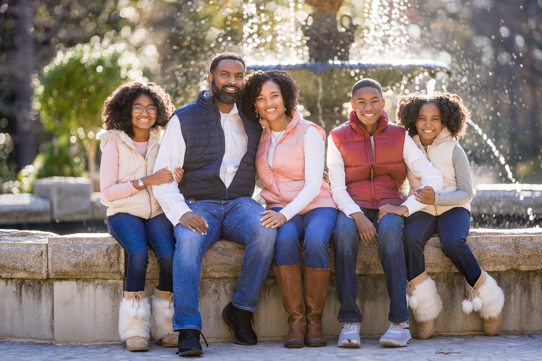 A family of five sits happily on a stone ledge in front of a fountain, capturing the joy of autumn mini sessions. They are all wearing winter attire, including vests and boots, against a backdrop of water spraying from the fountain and lush greenery.