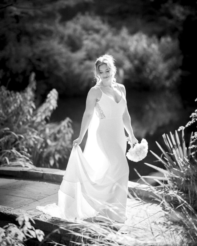 A bride in a flowing white dress stands outdoors near a pond, embracing her bouquet. The setting is lush with foliage at Duke Gardens, and she is backlit by soft sunlight, creating a serene, timeless scene perfect for photography.