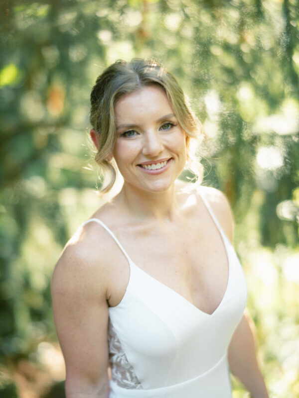 A smiling woman with light brown hair styled in loose waves is wearing a white dress. She stands outdoors at Duke Gardens, surrounded by blurred greenery and bathed in soft, natural light—a perfect setting for photography.