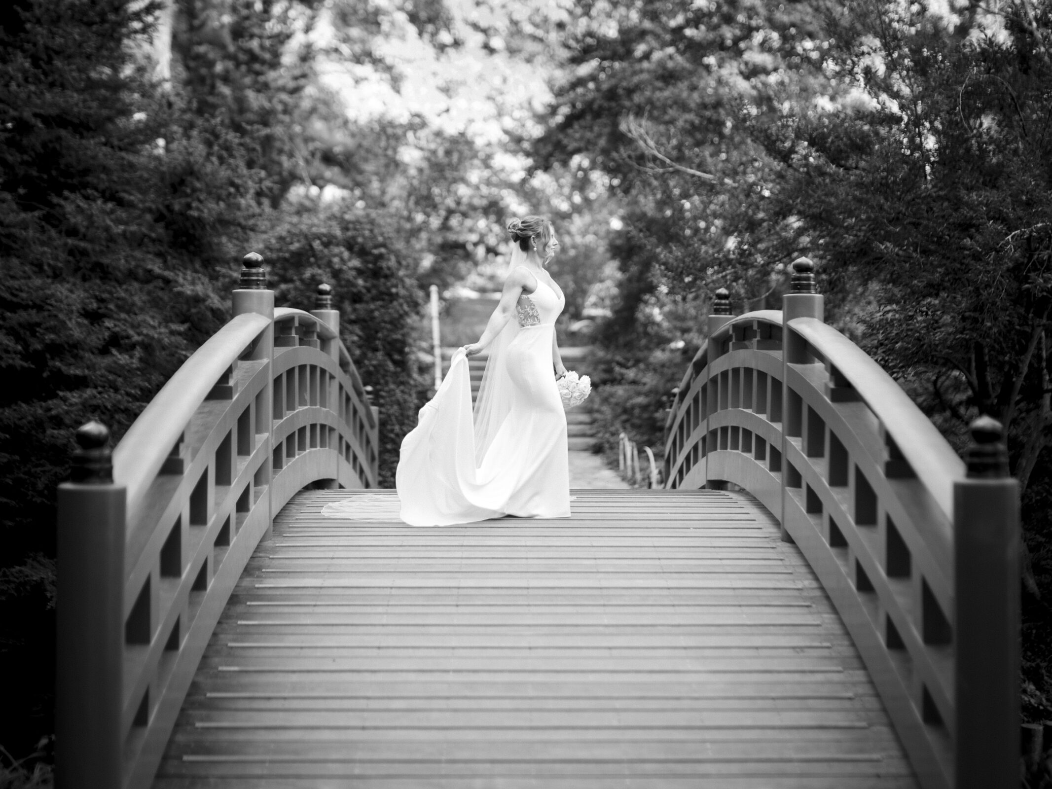 A black and white photo of a person in a flowing dress standing on an arched wooden bridge in Duke Gardens, surrounded by lush trees. The person is facing away, holding part of the dress, creating a serene and elegant scene captured with timeless photography.