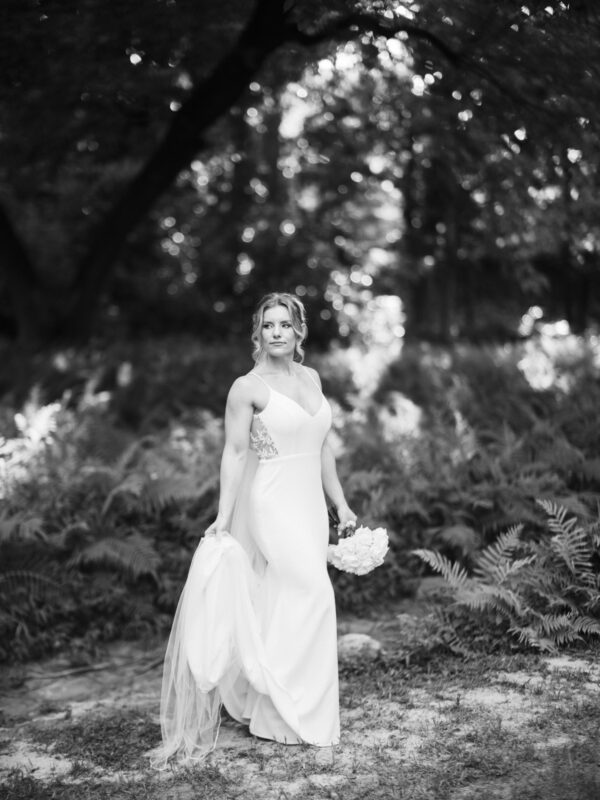 A woman in a white wedding dress stands in the wooded serenity of Duke Gardens, holding a bouquet and the train of her gown. Sunlight filters through the trees, casting soft shadows on the ground. The image is beautifully captured in black and white, showcasing timeless elegance.