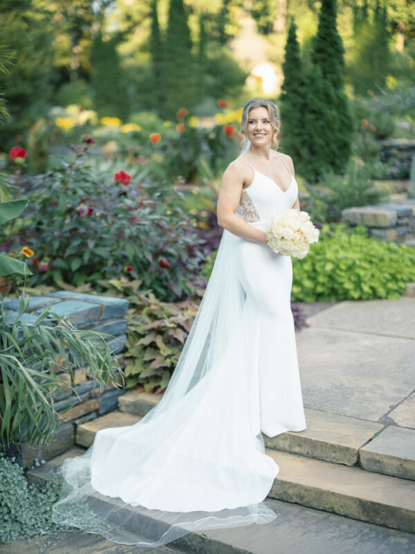 In the lush setting of Duke Gardens, a bride in a white wedding dress and veil holds a bouquet of white flowers on a stone path. She gazes joyfully to the side, surrounded by vibrant blooms and verdant greenery, capturing the essence of perfect garden photography.