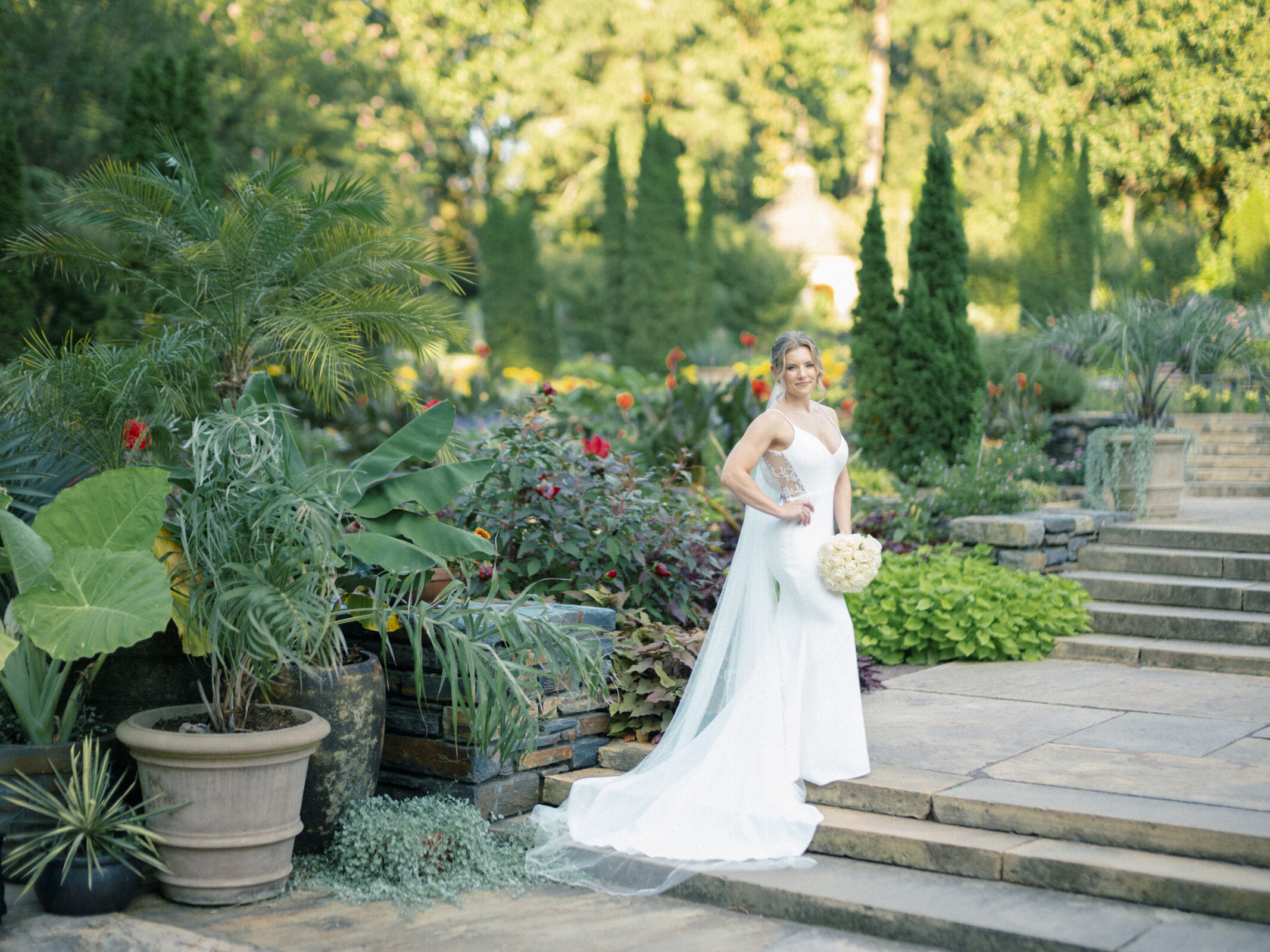 In the lush setting of Duke Gardens, a bride in a white gown poses gracefully, holding a bouquet. She stands on a stone patio amidst large potted plants and vibrant greenery, with steps ascending in the background under the sunlit sky—a perfect scene for timeless photography.