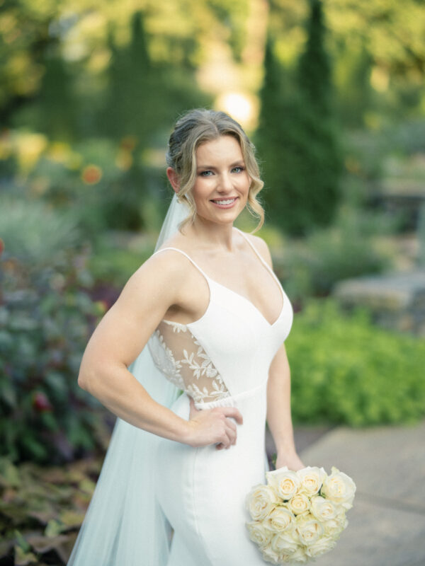 A smiling bride in a white dress with lace detailing holds a bouquet of pale yellow roses. She stands outdoors at Duke Gardens, surrounded by greenery and flowers, beautifully captured by natural light photography.