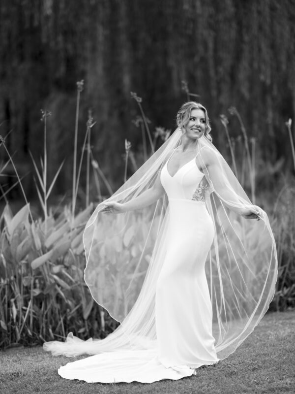 A bride in a long white dress and veil poses gracefully outdoors at Duke Gardens, smiling as she holds the edges of her veil. The slightly blurred background, with its tall plants, creates a soft and romantic atmosphere that's perfect for photography.