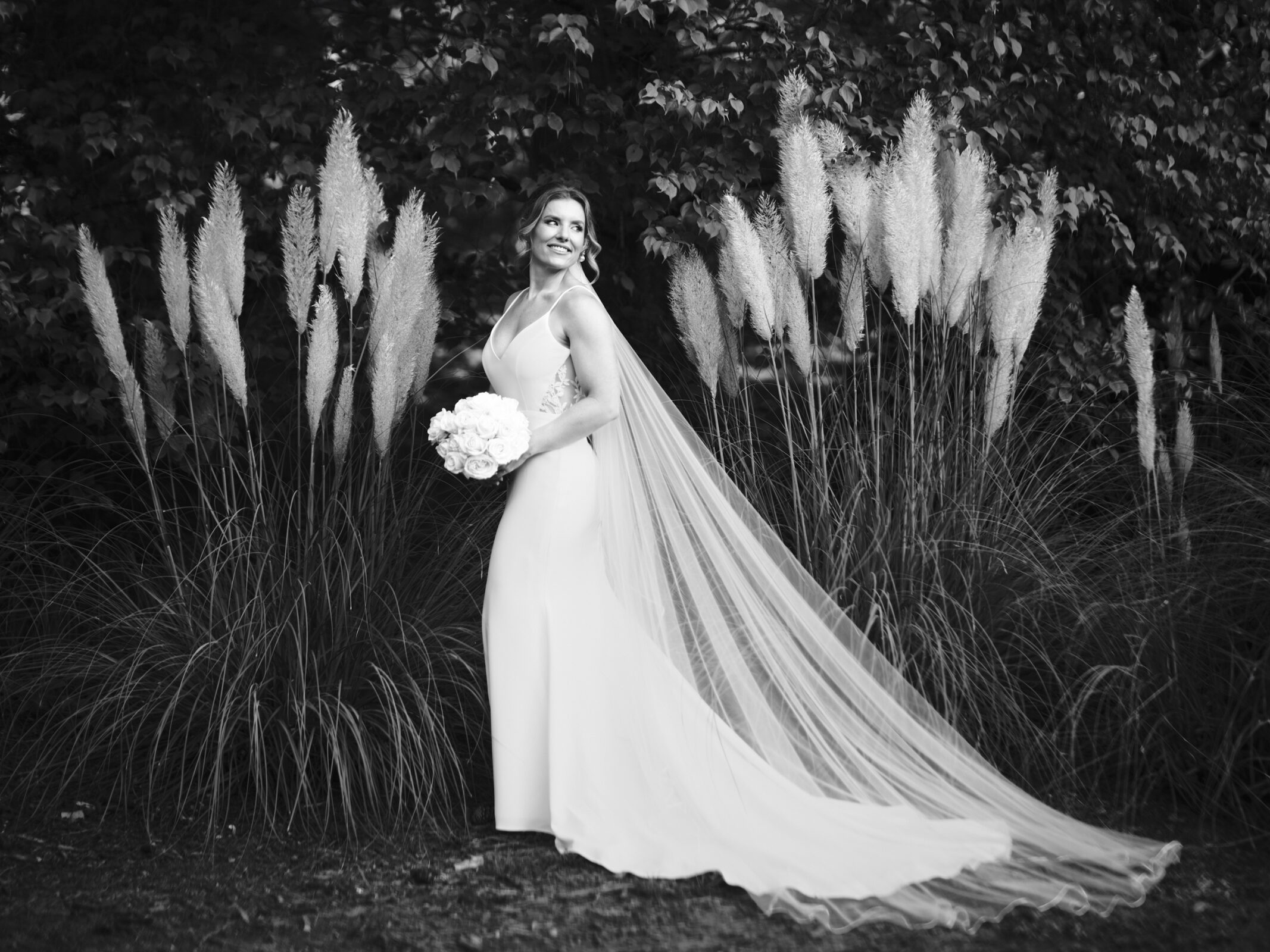 A bride in a flowing white gown and long veil stands holding a bouquet of flowers. She is smiling and looking to the side. Tall, feathery plants and dense foliage, reminiscent of Duke Gardens photography, form the background, creating a serene and elegant scene.