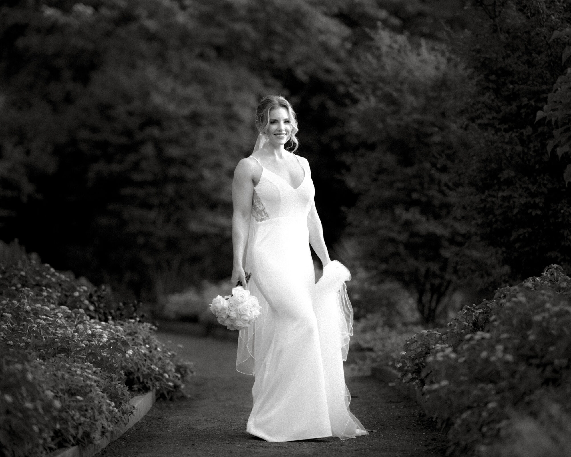 A bride in a long white dress holds a bouquet while walking along the garden path at Duke Gardens. The background features lush greenery, and the image is in black and white, highlighting the elegance of this iconic photography setting.