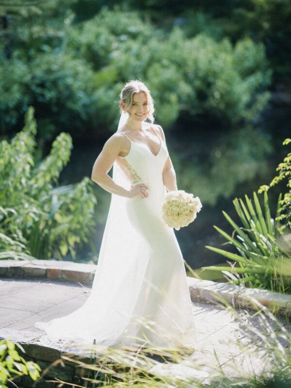 A bride in a white wedding gown stands outdoors on a stone pathway at Duke Gardens, holding a bouquet of white roses. The lush greenery and visible body of water create a serene and natural setting, perfect for photography.