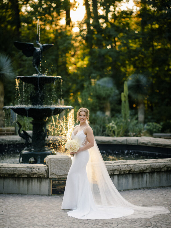 A bride in a white dress and long veil holds a bouquet of white flowers, captured perfectly by Duke Gardens Photography. She stands by a tiered fountain in the lush garden, with sunlight filtering through the trees in the background.