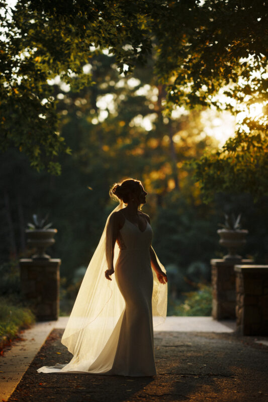 A bride in a flowing white dress stands outdoors under a canopy of trees, with sunlight filtering through the leaves, casting a warm glow. The serene scene at Duke Gardens Photography is picturesque, with lush greenery and stone pillars enhancing the enchanting background.