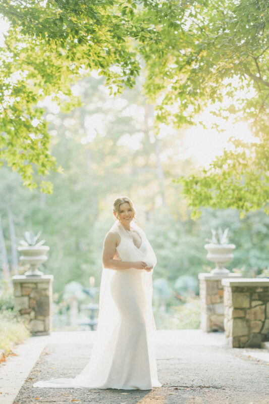 A bride in a white wedding dress stands on a path surrounded by greenery at Duke Gardens. She smiles with hands clasped, as sunlight filters through the trees, creating a warm and serene atmosphere. Two stone planters frame the picturesque scene, capturing perfect photography moments.