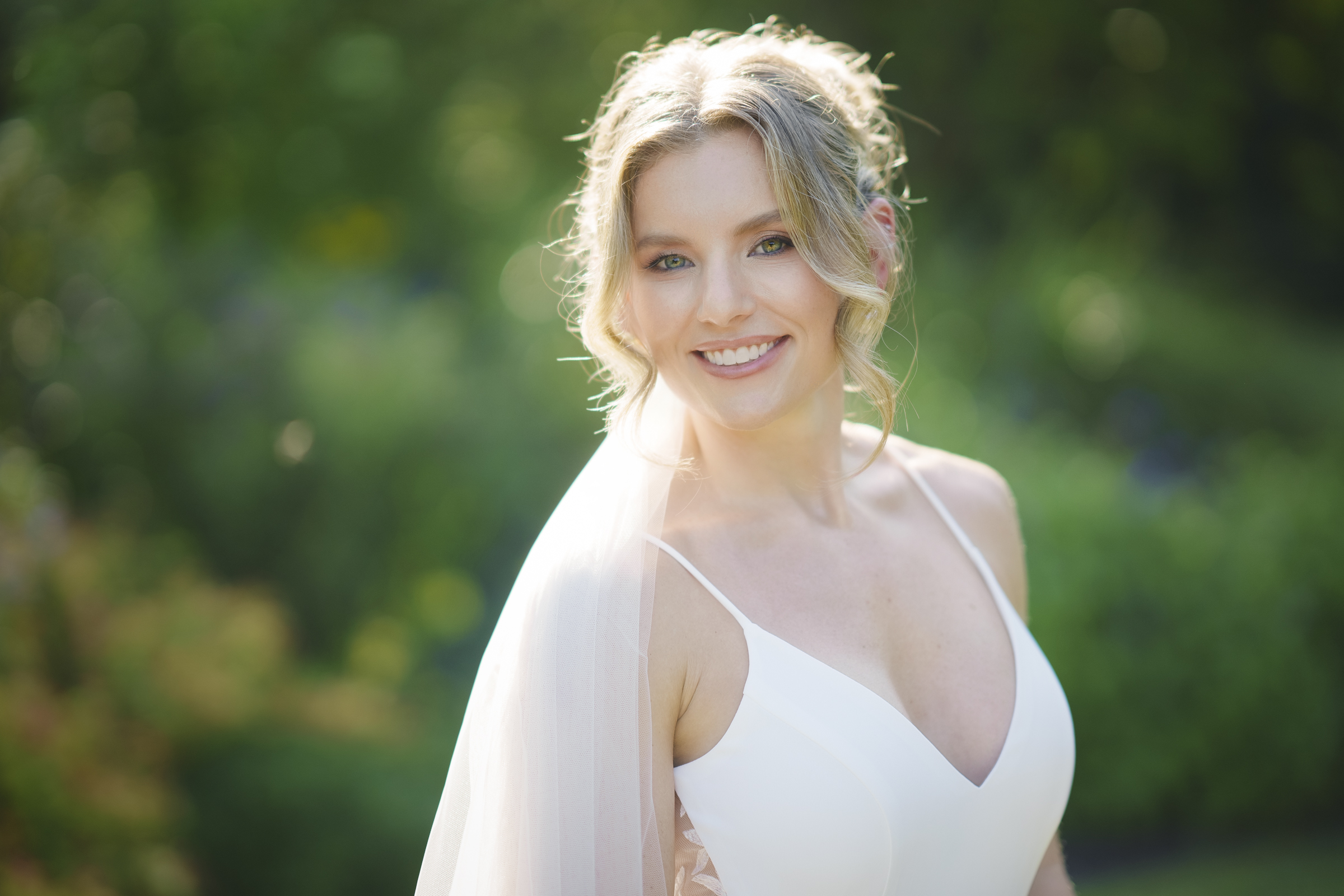 A woman in a white dress with a veil smiles brightly at the camera, capturing the elegance of Duke Gardens Photography. Her light hair is styled up, with loose curls framing her face against a lush, green outdoor setting softly blurred in the background.