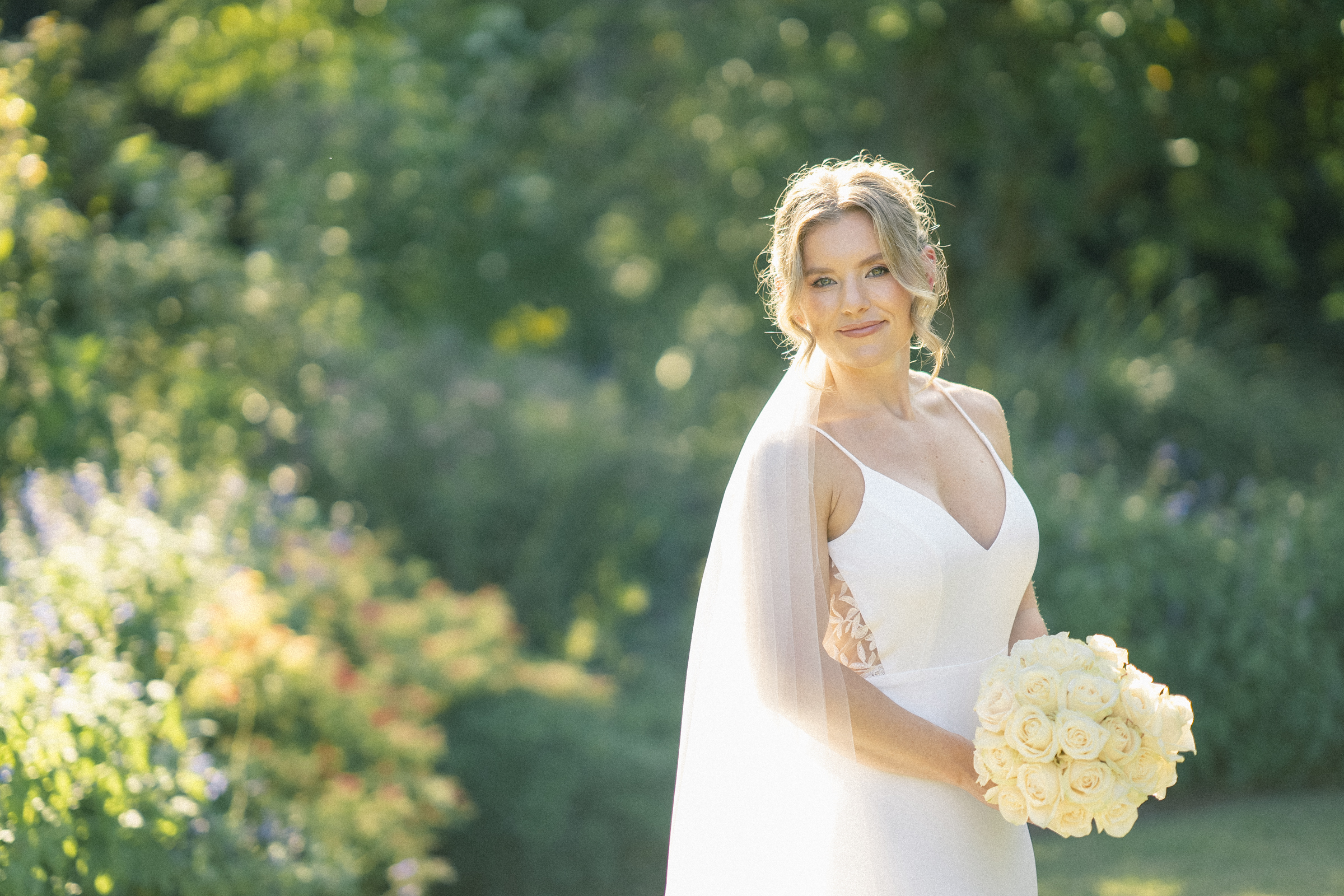 A bride in a white dress smiles while holding a bouquet of white roses. Captured by Duke Gardens Photography, she stands surrounded by lush greenery and colorful flowers, with sunlight filtering through the foliage.