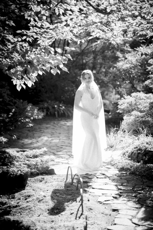 A bride in a white dress and veil stands on a sunlit stone path surrounded by lush foliage. The black and white photo, captured by Duke Gardens Photography, conveys the serene and timeless atmosphere of the garden setting.