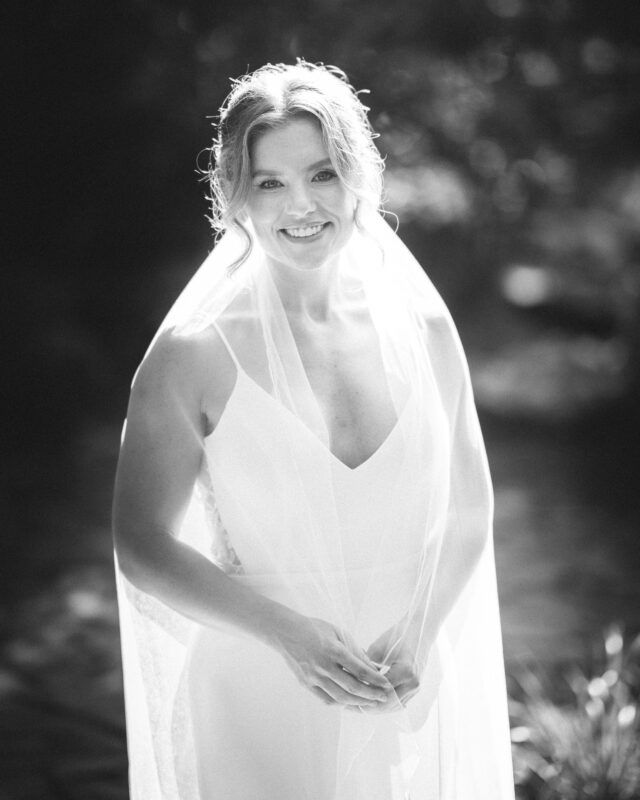 A smiling woman in a white dress and sheer veil poses outdoors, capturing the timeless elegance often celebrated in Duke Gardens Photography. The sunlight creates a soft, glowing effect around her, while the blurred hints of greenery enhance this black and white snapshot's ethereal charm.