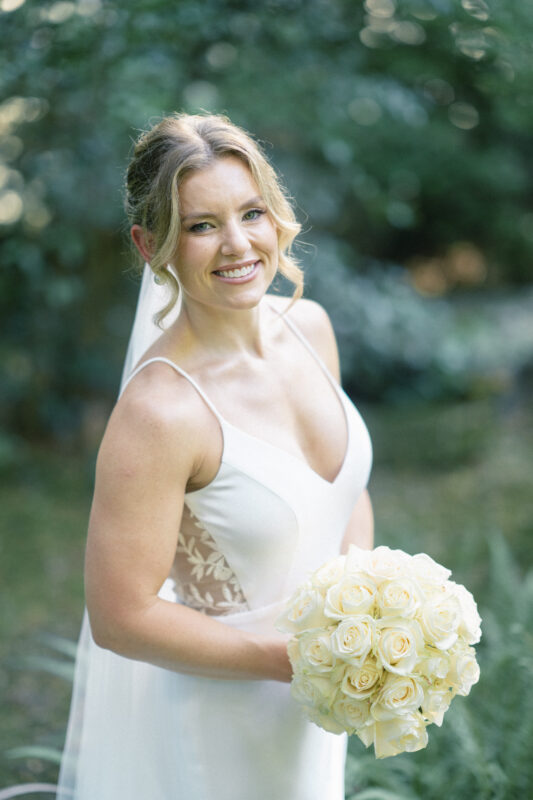 A smiling bride in a white wedding dress holds a bouquet of white roses. Captured by Duke Gardens Photography, she stands outdoors against a lush backdrop of greenery. Her hair is styled in an updo with loose curls framing her face, adding to the serene elegance of the moment.