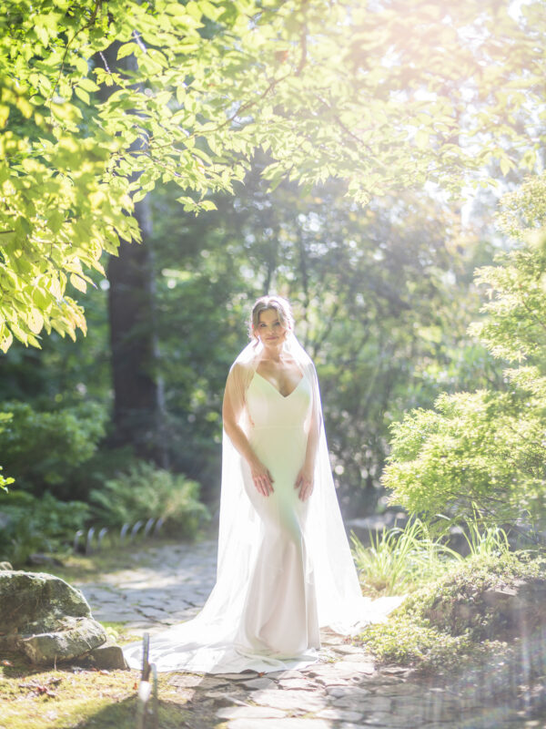 A bride in a flowing white gown stands on a sunlit path at Duke Gardens. Her long veil drapes around her shoulders as she gazes downwards, surrounded by vibrant green foliage and soft sunlight filtering through the leaves, perfectly captured by expert photography.