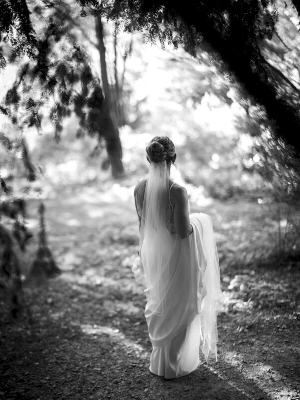 A bride stands on a forest path in Duke Gardens, wearing a long dress and veil. The black-and-white image captures the serene setting, with sunlight filtering through the trees, creating a peaceful and dreamy atmosphere akin to classic garden photography.