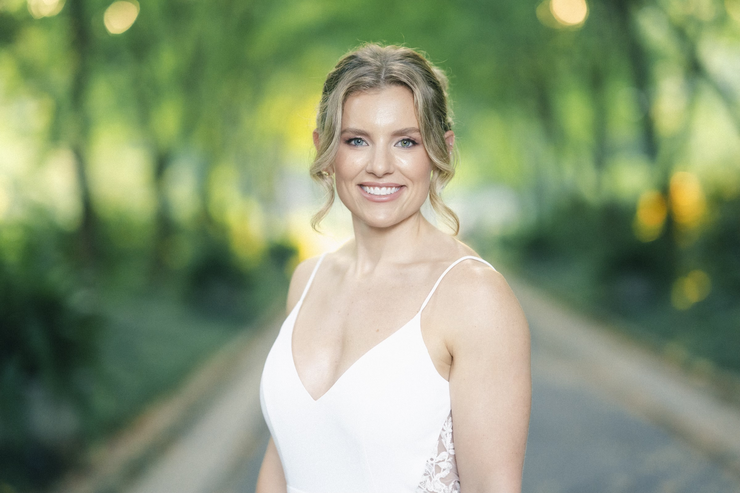 A smiling woman with light hair in an updo wears a white dress with lace details. She stands outdoors on a pathway surrounded by lush green trees, creating a serene and vibrant background that captures the essence of Duke Gardens photography.