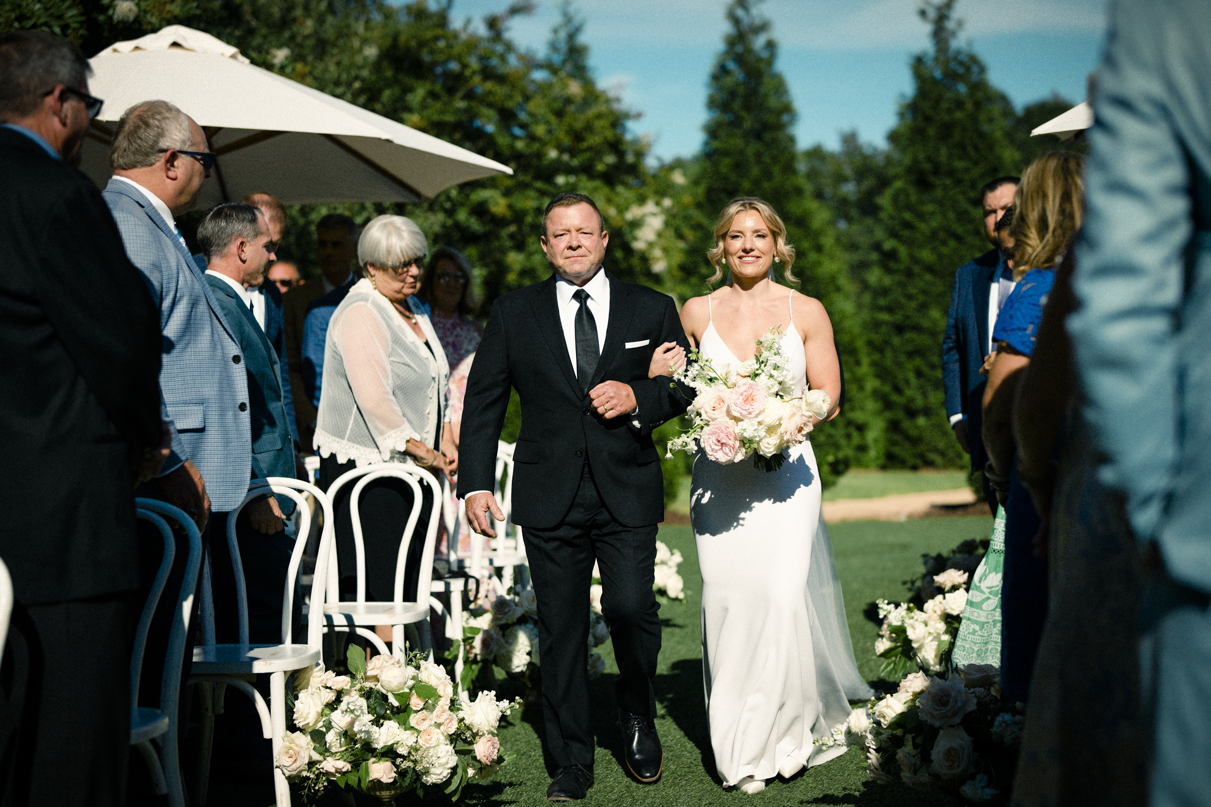 At The Bradford Wedding, a bride in a white dress gracefully walks down the outdoor aisle with a man in a suit. Guests seated on either side beneath white umbrellas are enveloped by lush greenery and vibrant flowers.