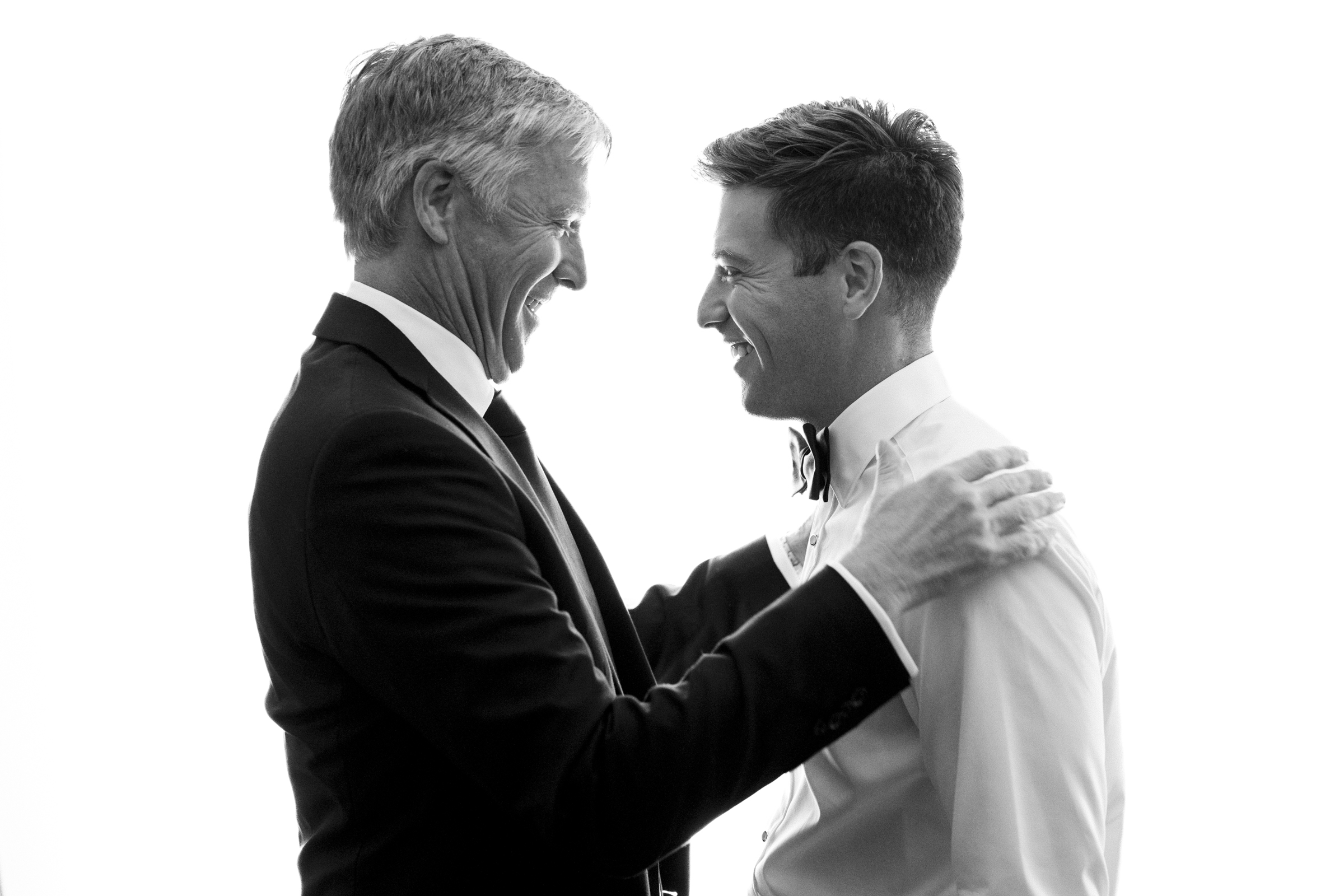 At The Bradford Wedding, two men in formal attire share a joyful moment, facing each other with wide smiles. The man on the left wears a dark suit, while the one on the right sports a light-colored shirt and bow tie. The background is bright and minimalist.