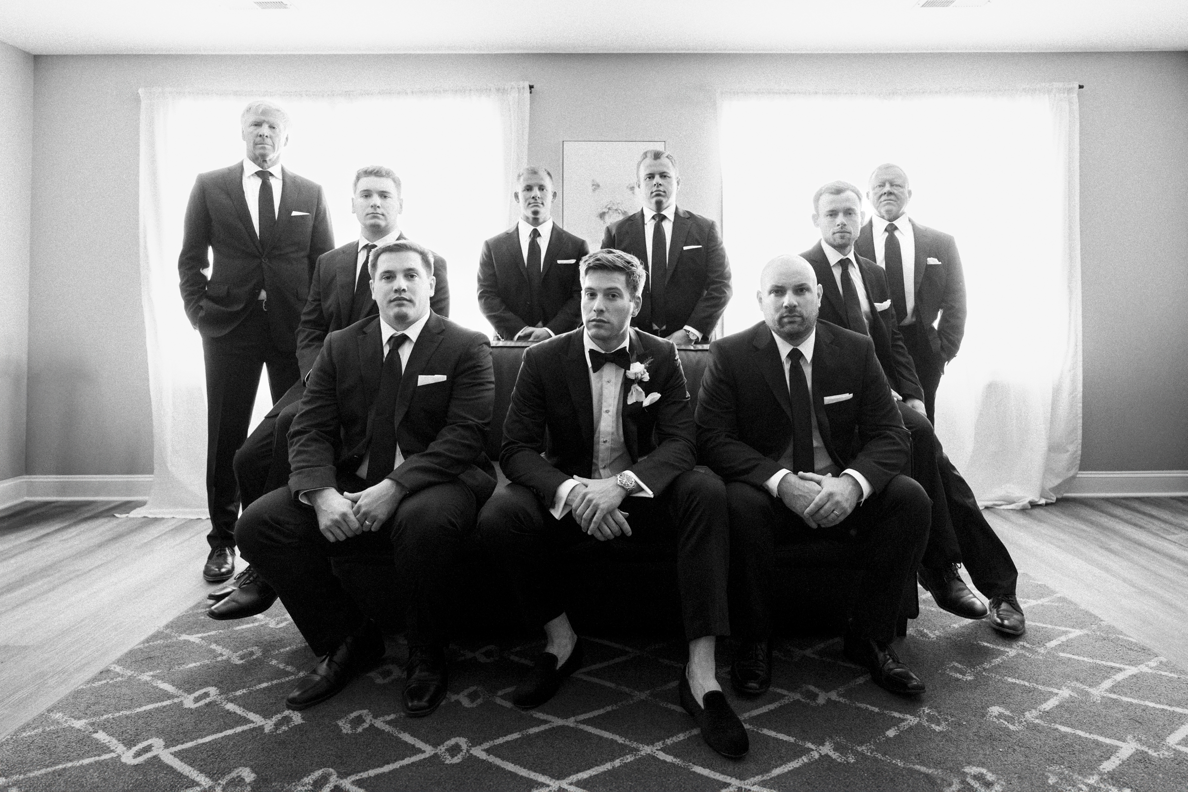 A black and white photo from The Bradford Wedding captures a groom and eight groomsmen in suits. The groom sits at the center, surrounded by his entourage in a room with large windows draped with sheer curtains, creating an atmosphere that's both formal and stylish.