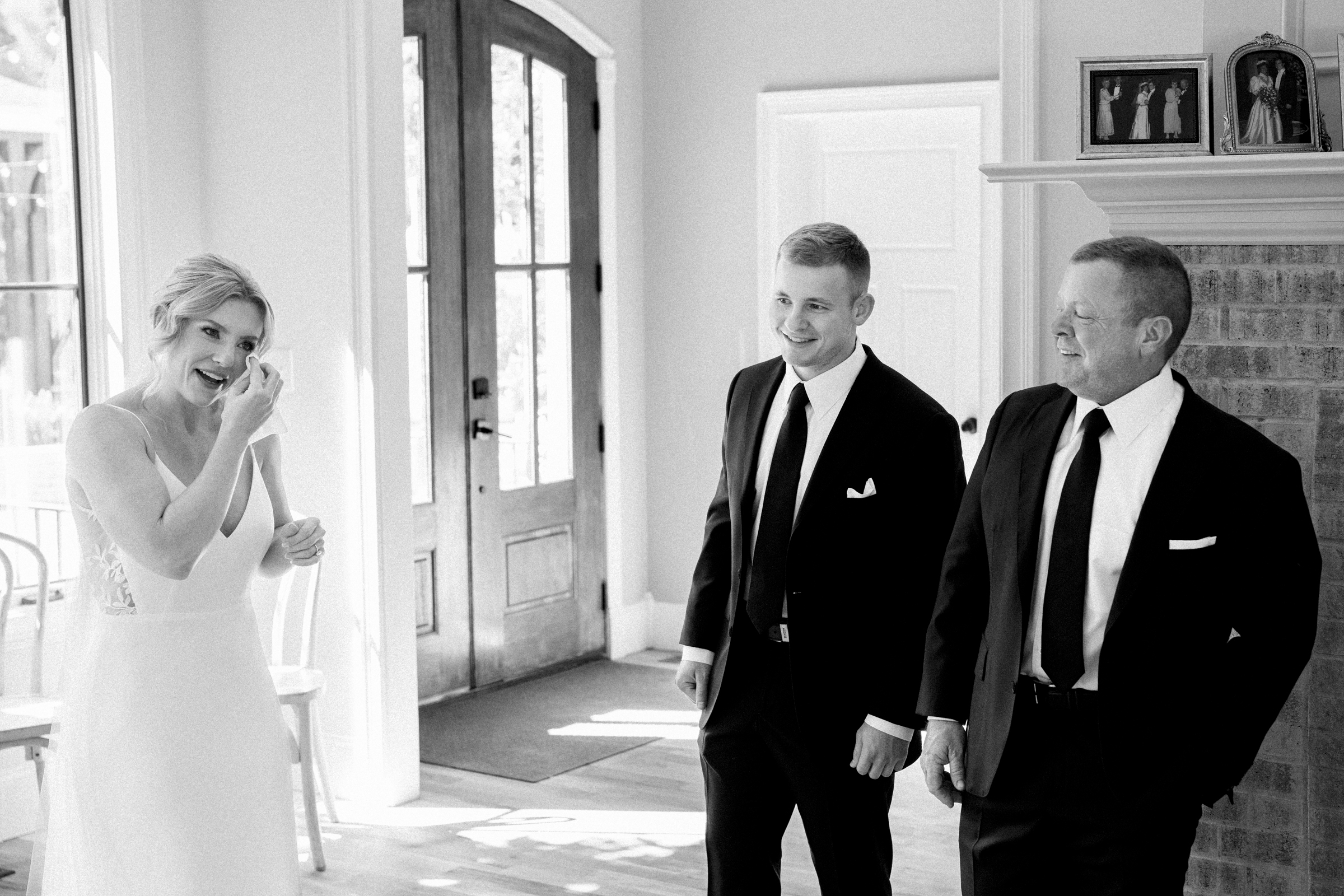 In a well-lit room with a brick fireplace and family photos on the mantel, a bride laughs joyfully while wiping a tear during the Bradford Wedding. Two men in suits, likely family members, stand nearby, smiling warmly at the touching moment.