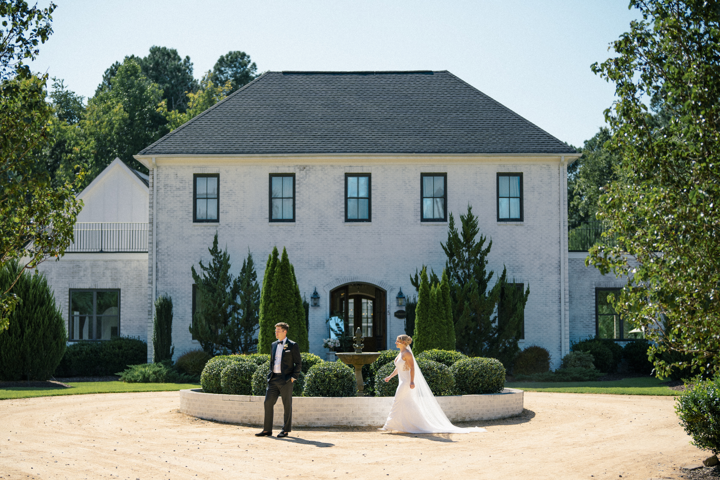 The bride and groom stand in front of a large, elegant white brick house with a circular driveway, embodying the timeless charm of The Bradford Wedding. The groom dons a black tuxedo, while the bride captivates in her flowing white gown. Trees and bushes frame them under the clear blue sky.