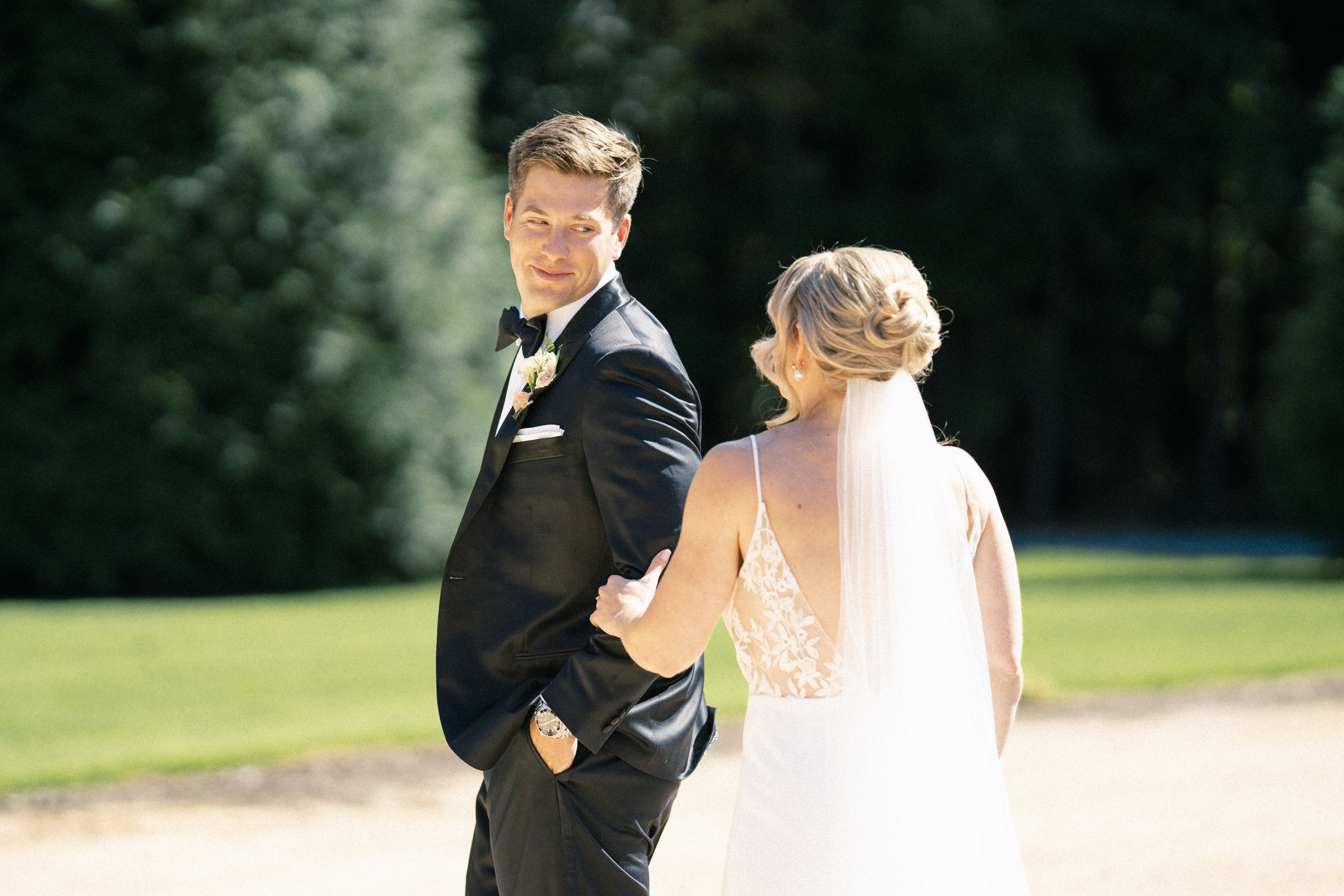 At The Bradford Wedding, a groom in a black tuxedo smiles at his bride in a white wedding dress and veil. They hold hands outdoors on a sunny day, surrounded by lush greenery.