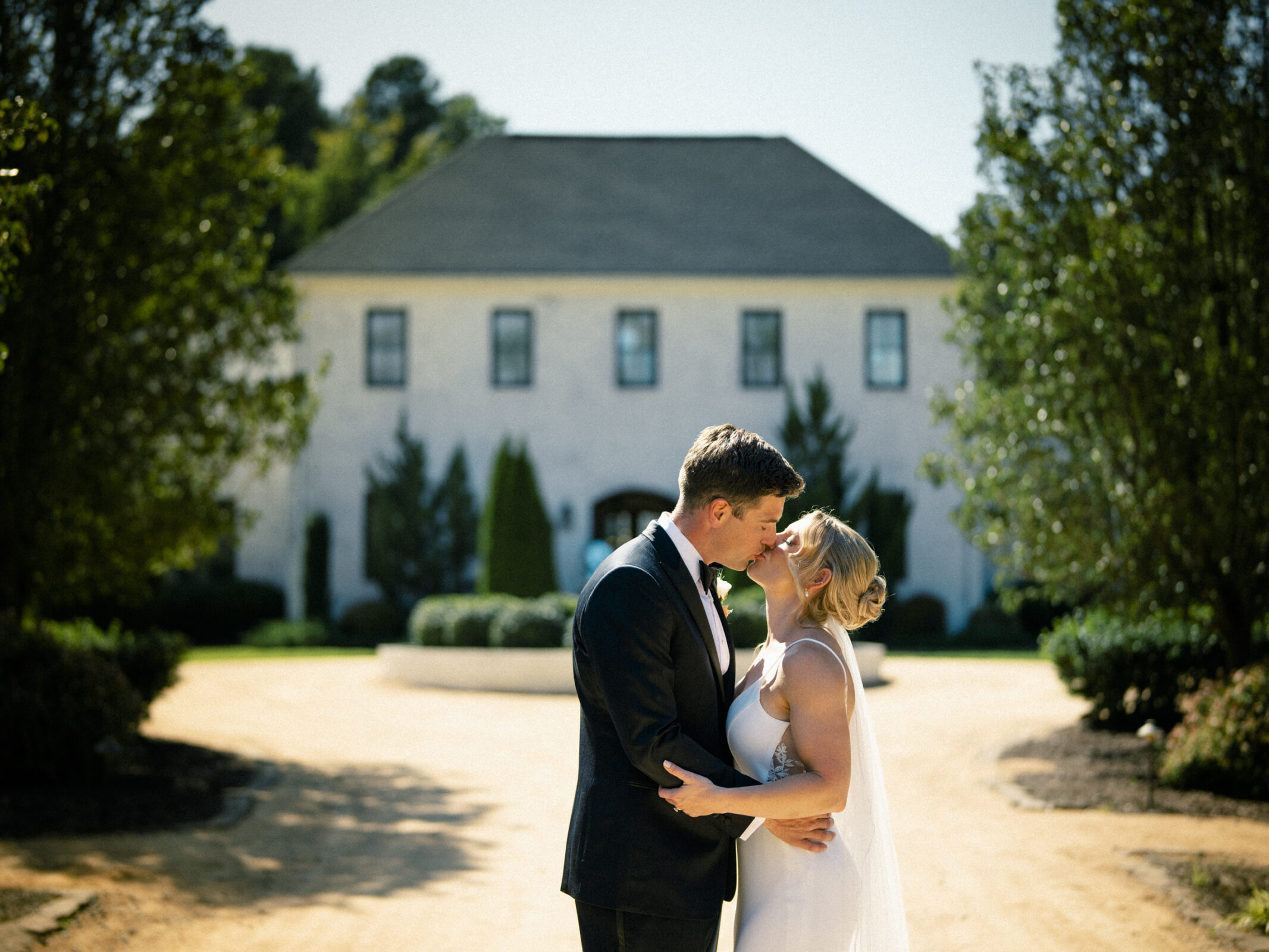 A couple dressed in wedding attire shares a tender moment outdoors during The Bradford Wedding. The groom in a black suit and the bride in a white gown stand close, with a large house and trees in the background, bathed in sunlight.