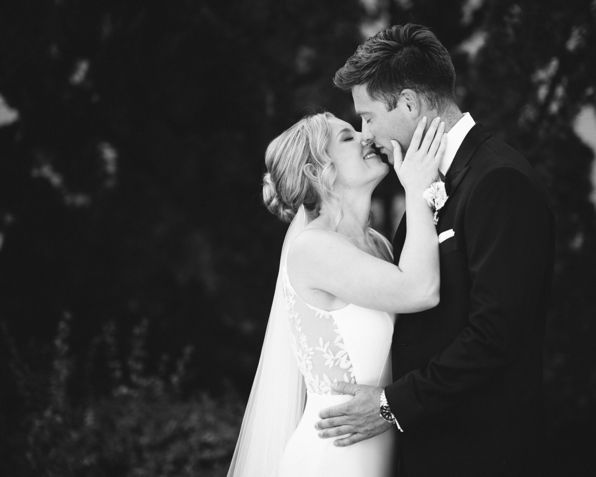 A black and white photo captures The Bradford Wedding, with a couple standing close together. The bride gently touches the groom's face, both smiling as the background softly blurs. She wears a veil and a dress adorned with lace details, encapsulating their timeless joy.