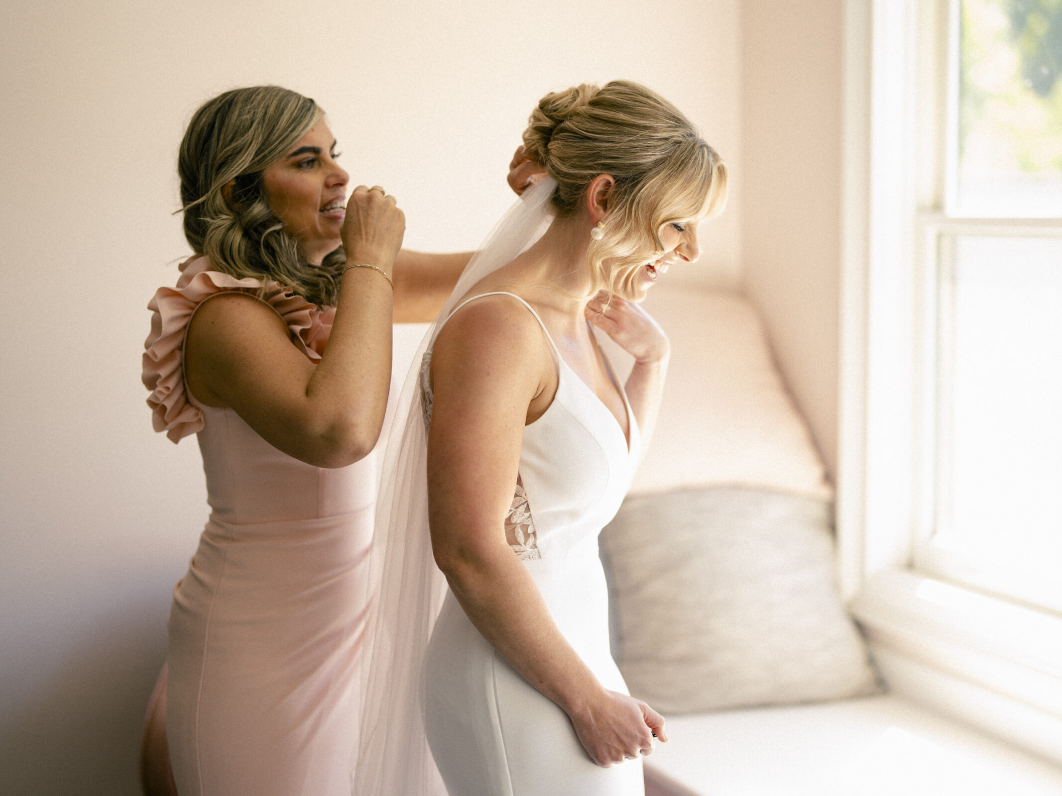 At The Bradford Wedding, a woman in a pink dress gently adjusts the bride’s veil by a window where soft light streams in, creating a warm and intimate atmosphere.