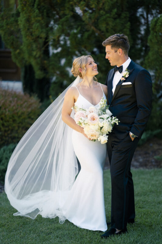 At The Bradford Wedding, a bride in a white dress and veil holds a bouquet of white and pink flowers, gazing lovingly at the groom in a black tuxedo. They stand outdoors on lush grass, surrounded by vibrant greenery, both smiling warmly at each other.