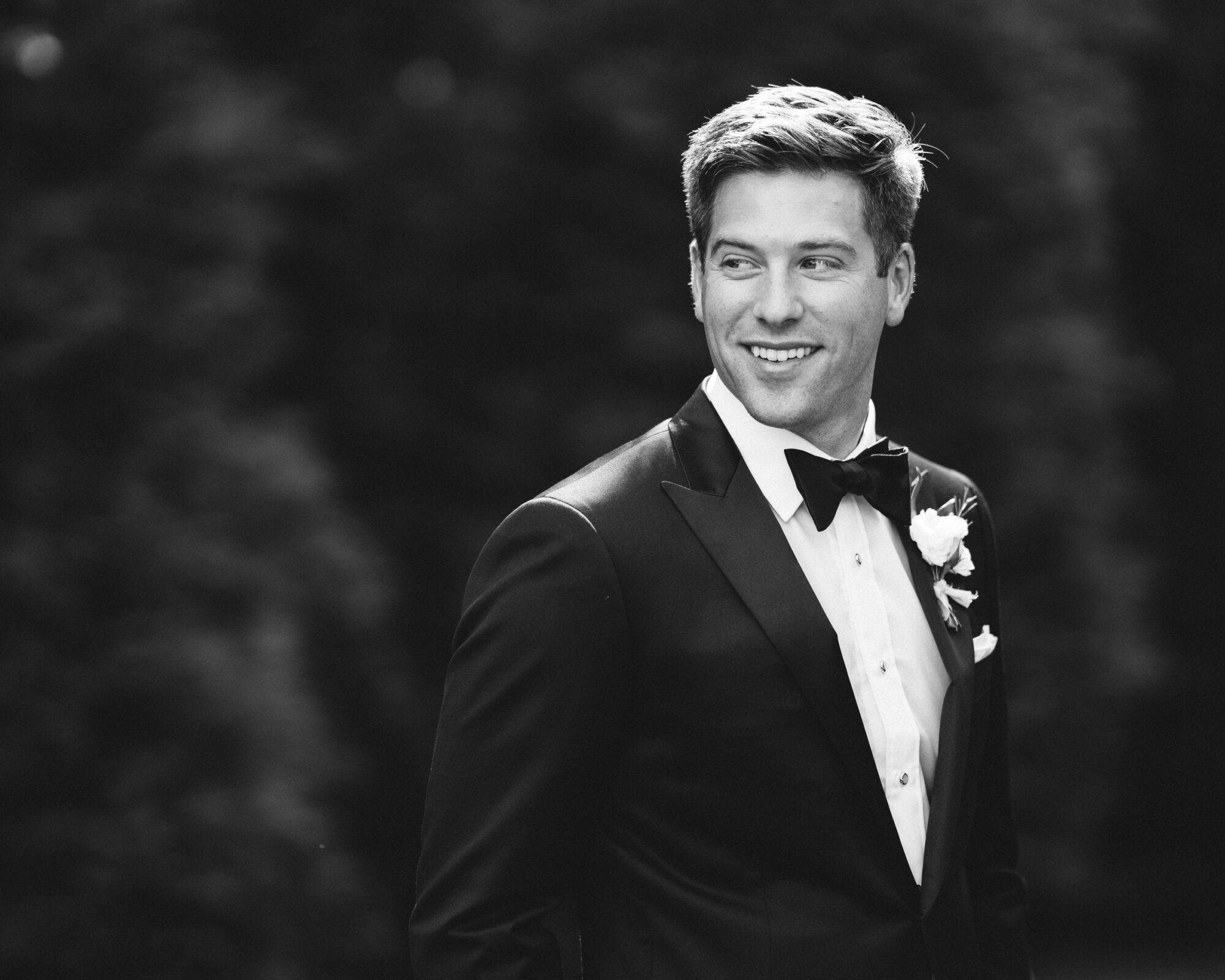 A black and white photo captures a man in a tuxedo, bow tie, and flower boutonniere, smiling as he gazes to his left. The background blurs into dark foliage, preserving the elegance of The Bradford Wedding.