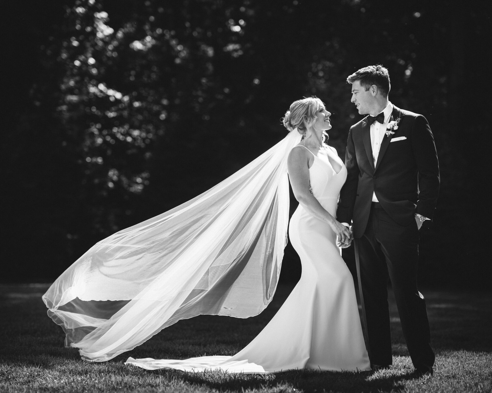 In an elegant black-and-white photograph, The Bradford Wedding captures a bride in a flowing dress with a long veil and a groom in a tuxedo. They stand outdoors, holding hands and gazing into each other's eyes.