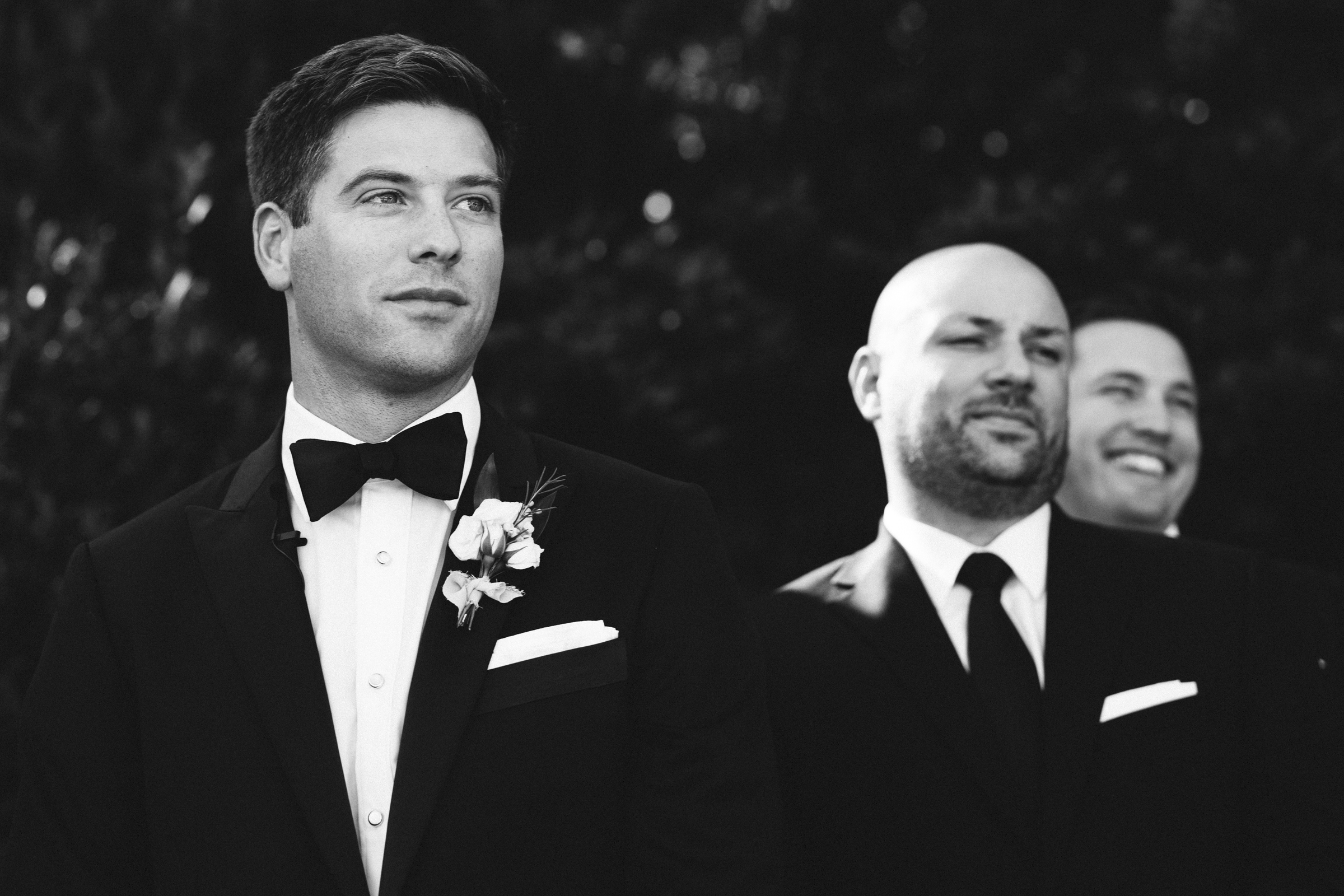 Black and white photo of three men in suits. At The Bradford Wedding, the man in the front, wearing a bow tie and floral boutonniere, gazes thoughtfully to the side. Behind him, two men smile in suits and ties, enhancing the sense of camaraderie.