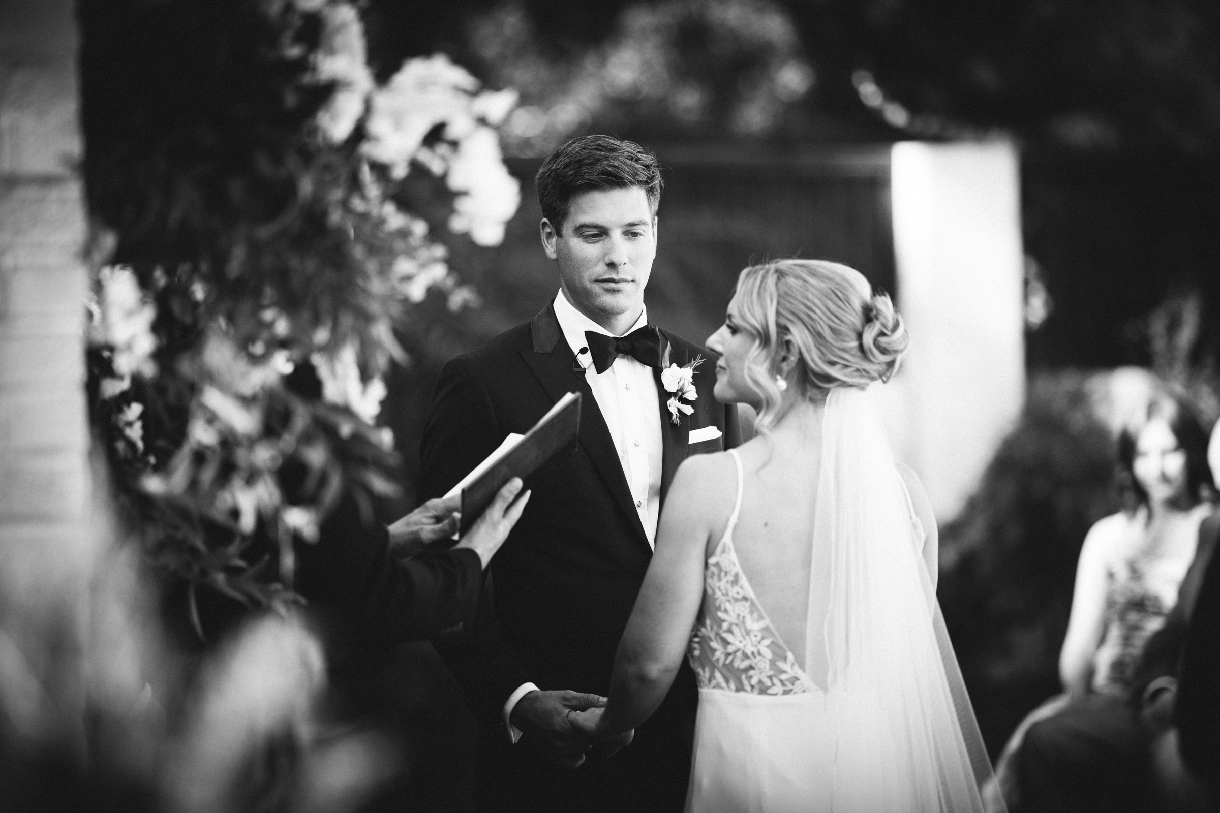 In the timeless black and white scene of The Bradford Wedding, a groom in a tuxedo and a bride in a veil hold hands, gazing into each other's eyes. A person with a book stands before them, likely officiating, while guests linger softly blurred in the background.