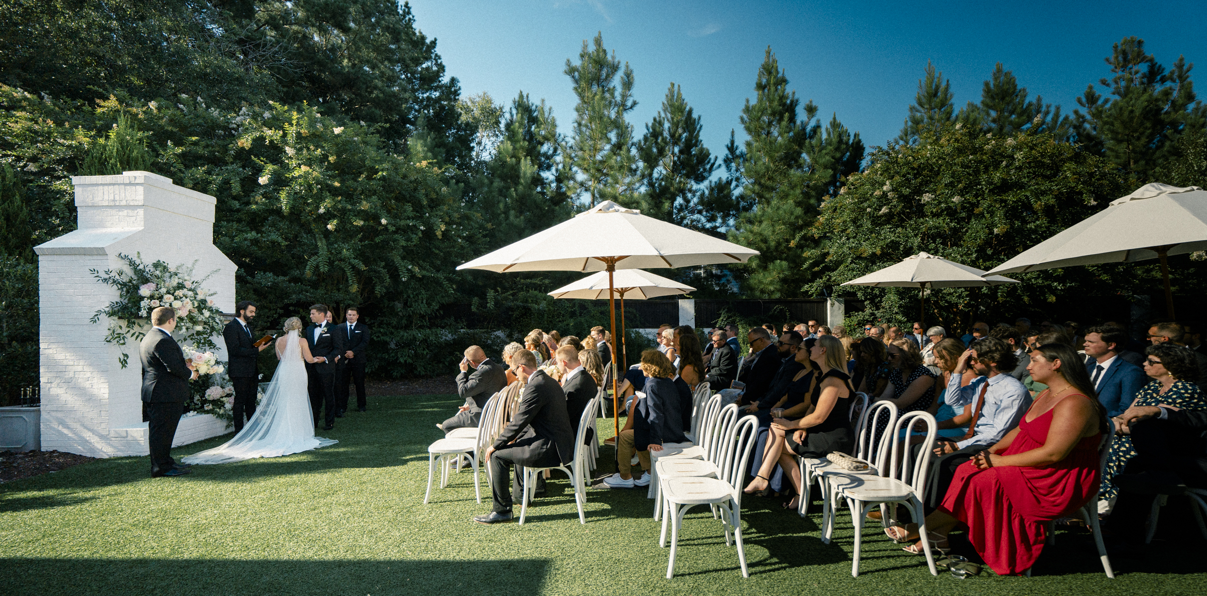 At The Bradford Wedding, an outdoor ceremony unfolds with a couple standing by a white floral arch. Guests are seated on white chairs under umbrellas, surrounded by trees. The sun shines in a clear blue sky, creating a serene and warm atmosphere.