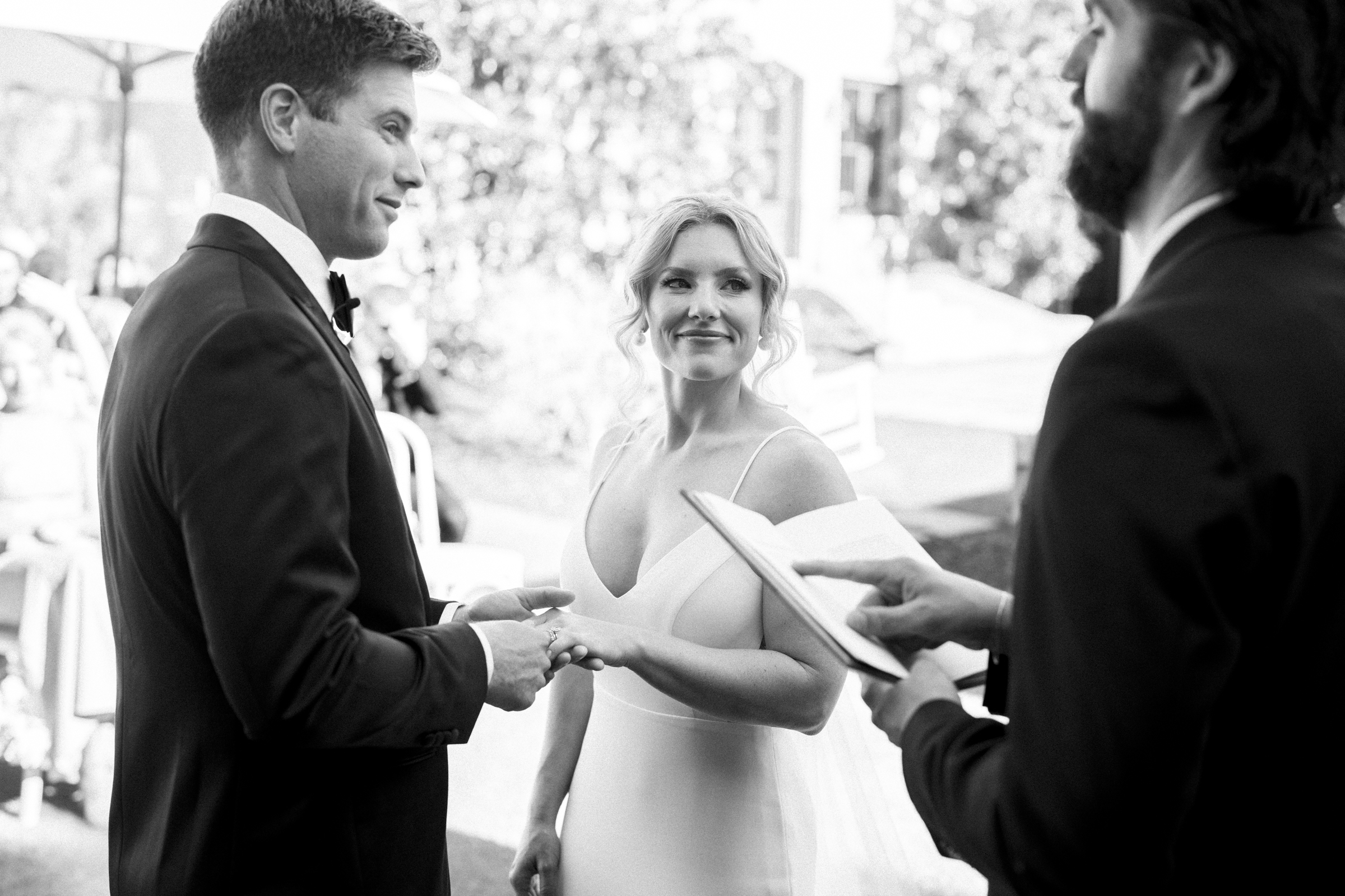 During The Bradford Wedding, a bride and groom exchange rings in an outdoor ceremony, with a suited officiant holding a book in the foreground. The bride smiles warmly at the groom, who is dressed in a tuxedo. The setting is bright and joyful.