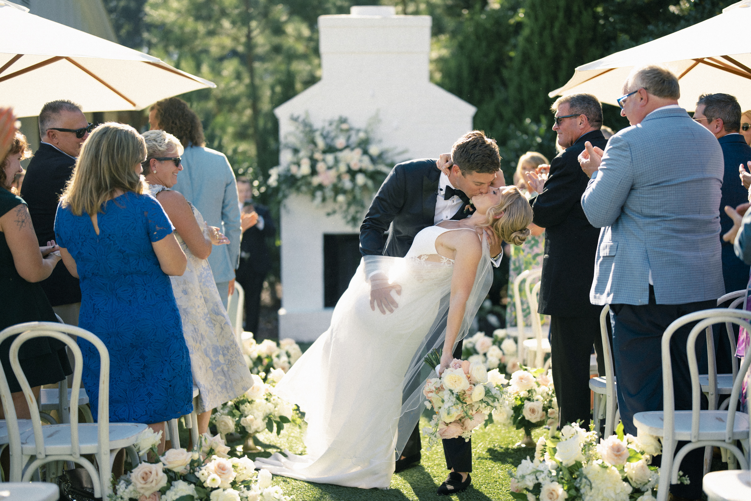 At The Bradford Wedding, a bride and groom share a kiss on a sunlit, flower-lined aisle. Guests in formal attire applaud from either side, some sheltered under umbrellas. In the background, a white fireplace adorned with blooms adds charm to this elegant outdoor celebration.