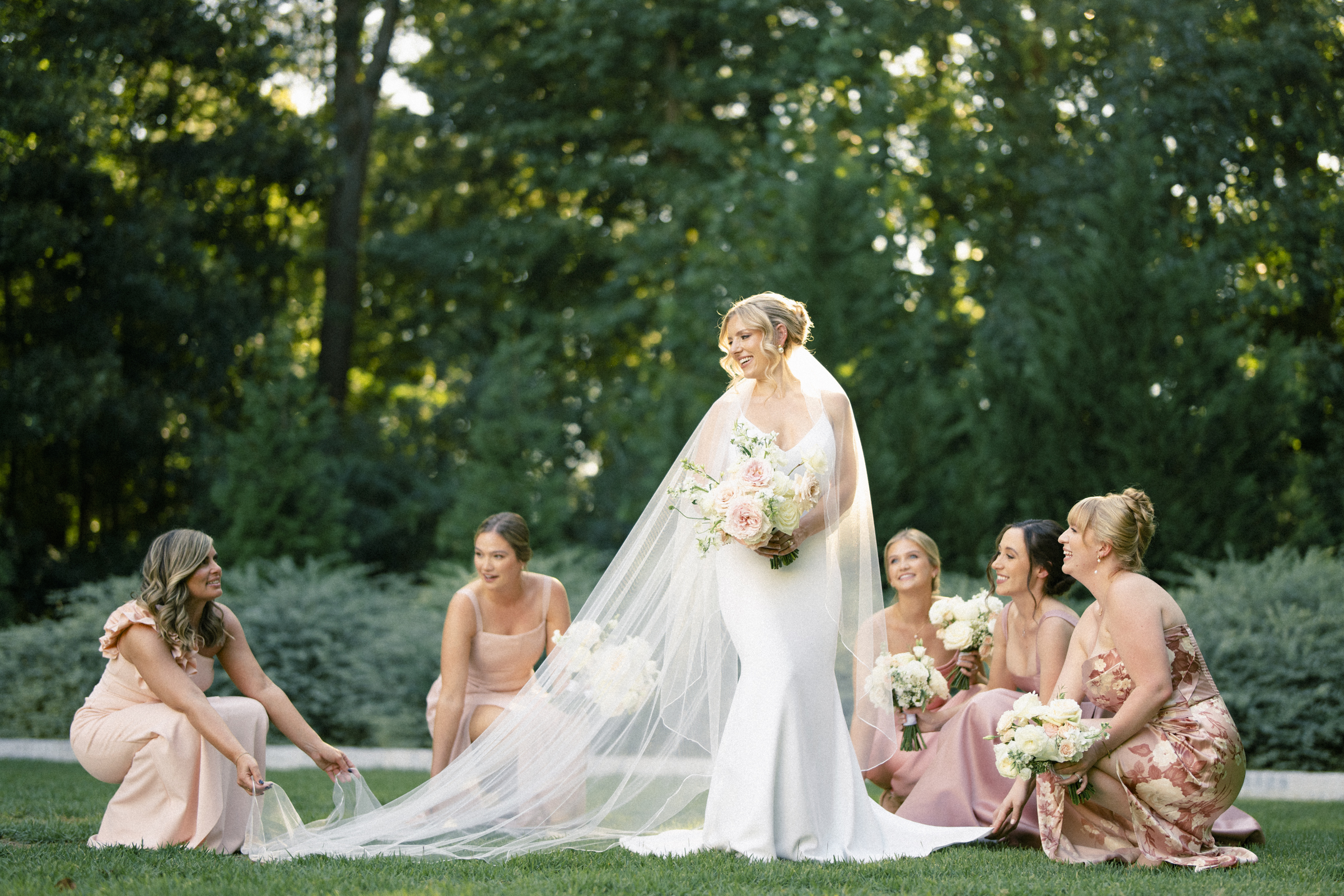 At The Bradford Wedding, a bride in a white dress and long veil stands smiling with a bouquet, surrounded by five bridesmaids in blush dresses. On the lawn, framed by trees, two bridesmaids gently adjust her veil as the scene unfolds beautifully.