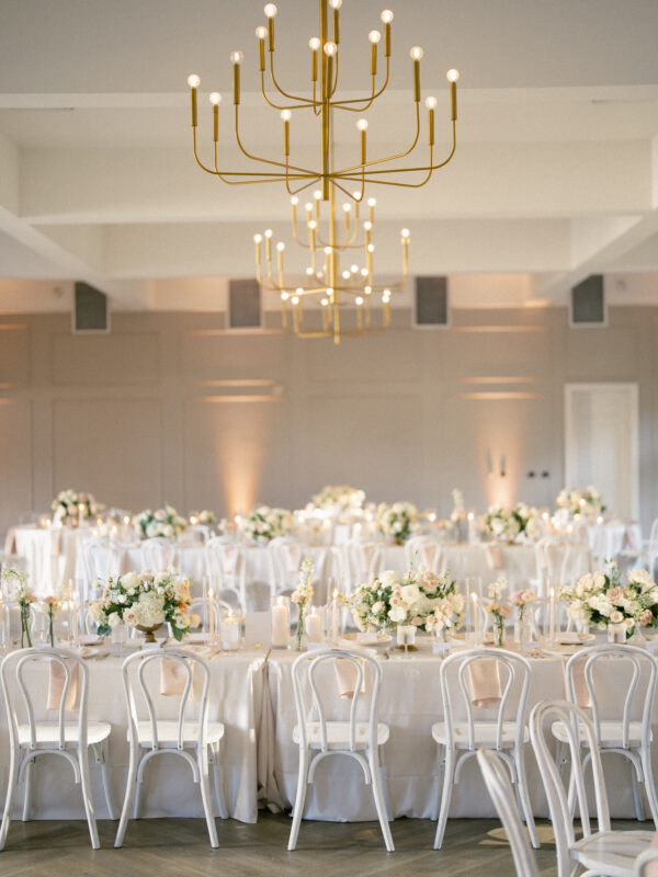 The Bradford Wedding features a beautifully decorated reception hall with elegantly set tables adorned with white linens and floral centerpieces. Surrounded by white chairs, the tables bask in the warm glow of modern chandeliers hanging from the ceiling.