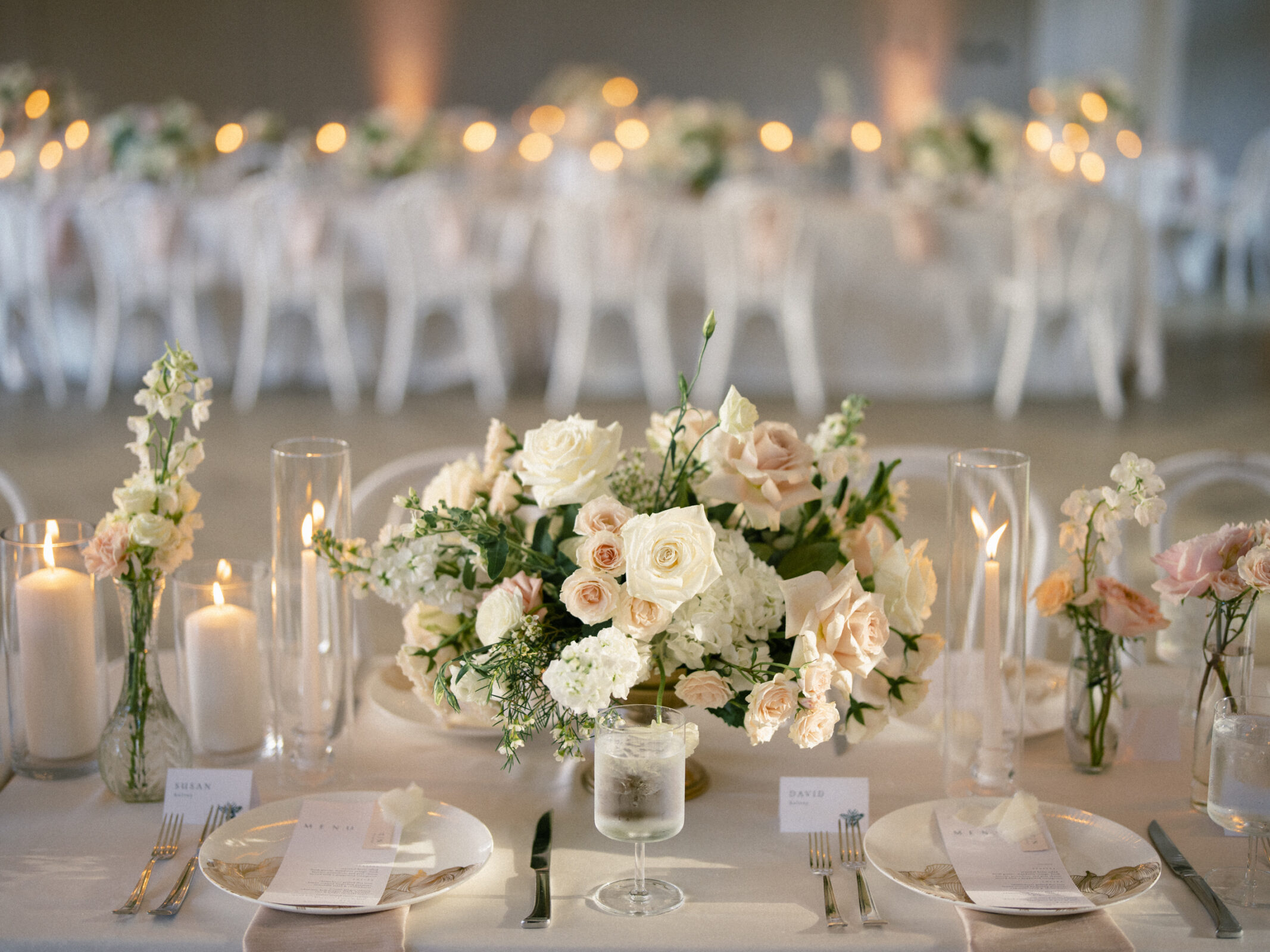 The Bradford Wedding featured an elegant reception table setting with a pastel floral centerpiece surrounded by lit candles. Plates with menus and cutlery were neatly arranged. A softly lit background with more decorated tables enhanced the romantic ambiance.