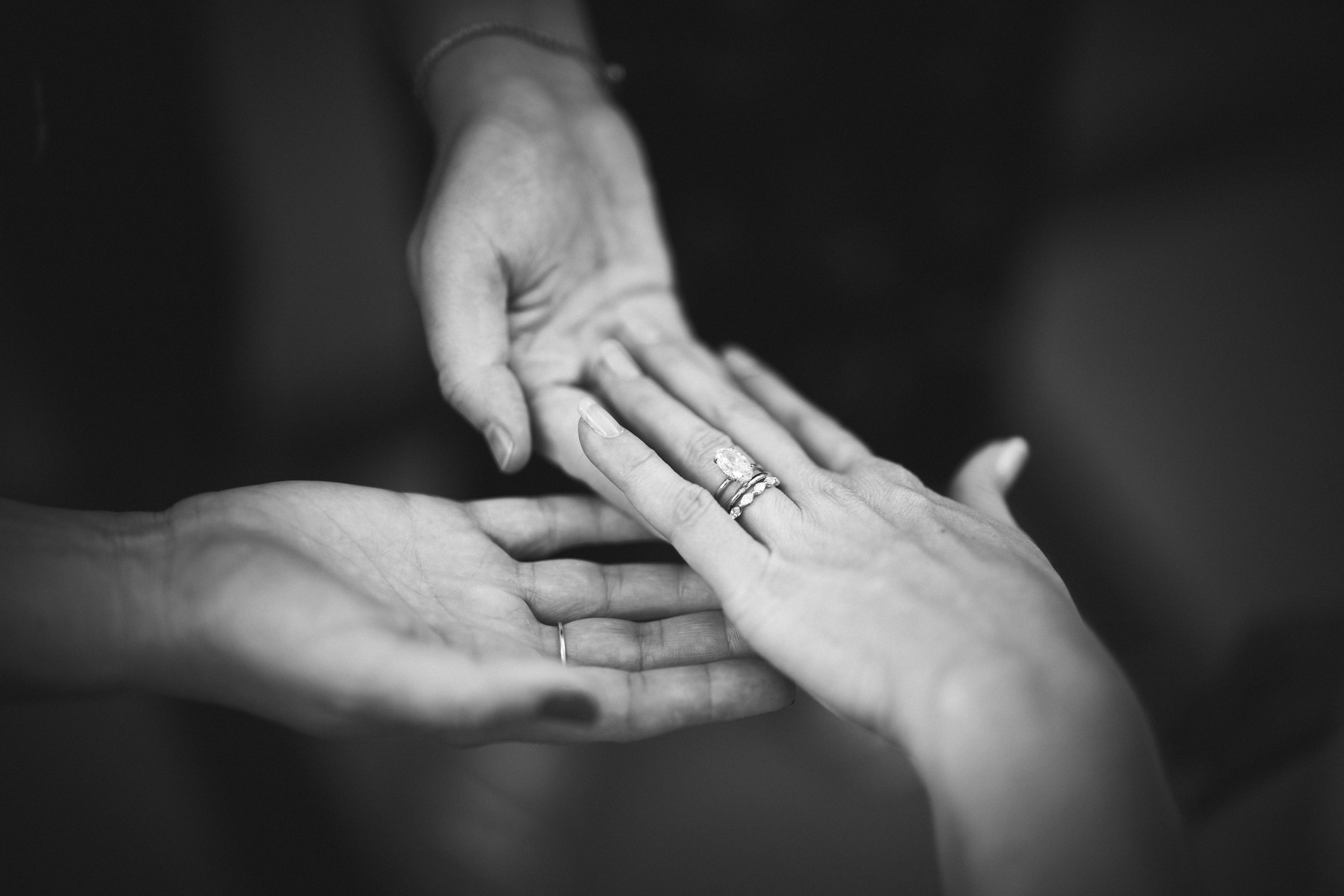 A black and white image captures two people at The Bradford Wedding, holding hands. One hand showcases a ring, while the other gently supports it from below. The focus on their hands suggests a moment of profound connection and support.