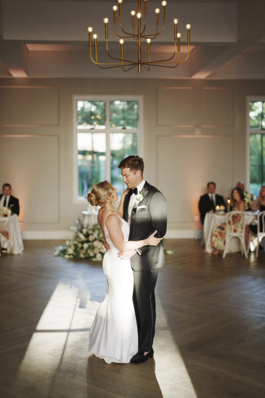 At The Bradford Wedding, a bride and groom share their first dance in an elegant reception hall with soft lighting. She wears a white dress, he a black suit. Guests sit at tables adorned with candles and flowers, while sunlight streams through windows beneath a grand chandelier.